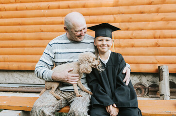 senior elderly man grandfather holds poodle dog in his arms and hugs his grandson boy who graduate...