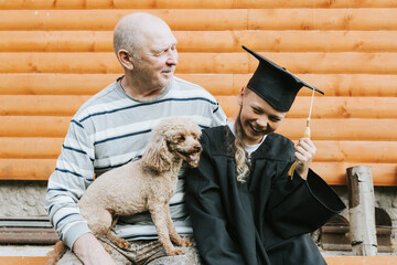 senior elderly man grandfather holds poodle dog in his arms and hugs his grandson boy who graduate...