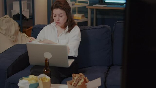 Young woman using laptop on sofa while eating asian food with chopsticks. Caucasian person sitting at home with chinese takeaway meal, looking at movie on TV and working on gadget