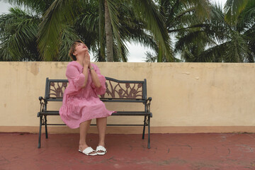 An asian woman desperately prays for a miracle while looking up to the heavens. A lady in her 20s sitting on a bench praying.