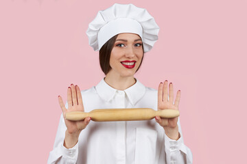 A young girl cook in a white robe and a cap stands and holds a rolling pin for cooking isolated on a pink background.