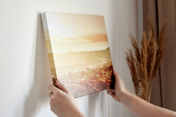Canvas print with gallery wrap. Woman hangs landscape photography on white wall. Hands holding...