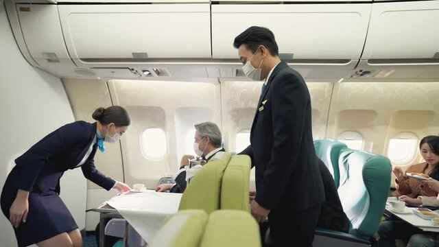 Caucasian female and Asian male flight attendants with trolley cart, serving a cup of coffee to a middle-aged Caucasian male passenger on seat. Three of them wearing face masks for protective Covid-19