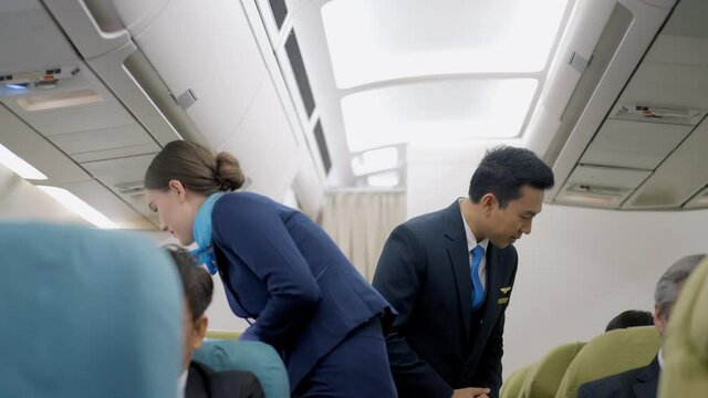 Caucasian female and Asian male flight attendants in uniforms walk along the aisle checking the passengers on safety standards with seat belt fastened, upright seat backs before airplane take off.