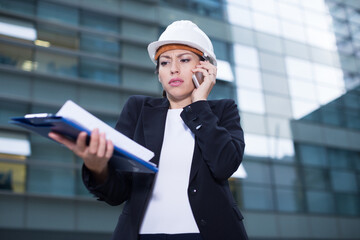 Young woman engineer in suit and hat is taking folder and discussing project by the phone near the building.