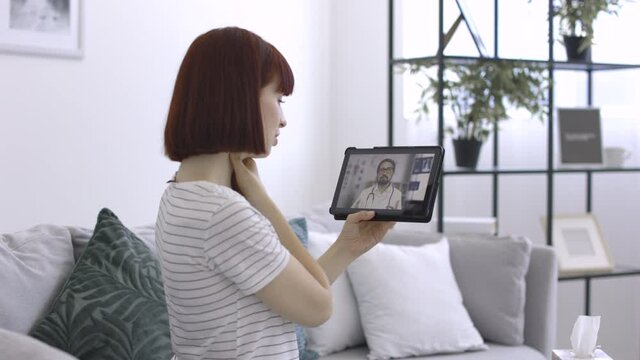 Young sick woman in casual wear, sitting on sofa at home and talking online with her physician using tablet computer about her symptoms, sore throat, cough and blocked nose. Online appointment