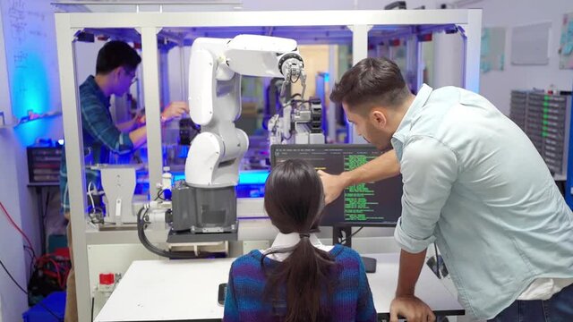 Back of an Asian female apprentice engineer sitting at a desk keying a robotic software code on a computer, with a Caucasian male engineer standing beside her to give advice in an industrial factory.