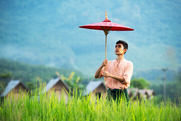 Burmese man walking holding a red umbrella In the rice field behind is an old house. Myanmar space for text