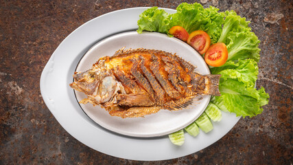 tasty large fried nile tilapia fish with lettuce, tomato and cucumber in ceramic plate on rusty texture background, top view