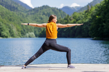 Fototapeta na wymiar Young woman doing warrior yoga pose near the lake.