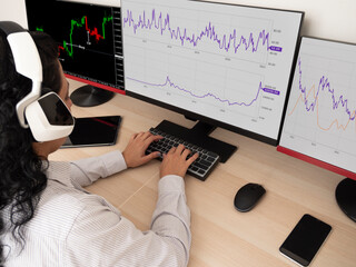 Caucasian young woman sitting at a table doing stock trading on a computer with three black monitors in a white room