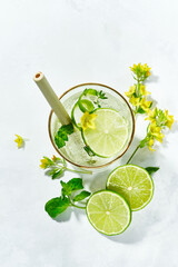 Close-up of the top of a glass with a garnishing of fresh flowers, lime slices inside summer detox refreshing drinks on white background. Sunny daytime shadows from a mocktail.