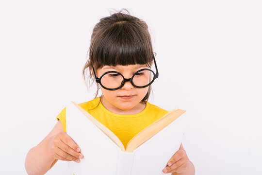 Smiling little girl, wearing yellow t-shirt and round black glasses holding an open book in her hands and reading, on white background