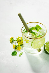 Close-up of the top of a glass with a garnishing of fresh flowers, lime slices inside summer detox refreshing drinks on white background. Sunny daytime shadows from a mocktail.