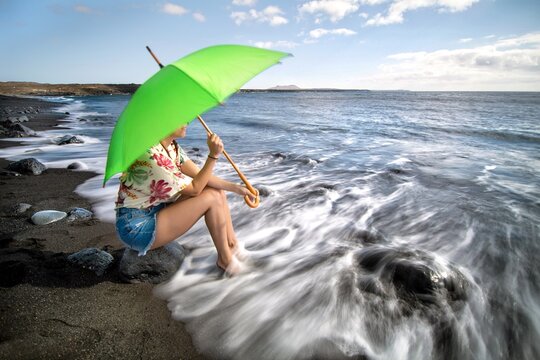 Joven muchacha con paragua verde sentada frente al mar en una playa de rocas