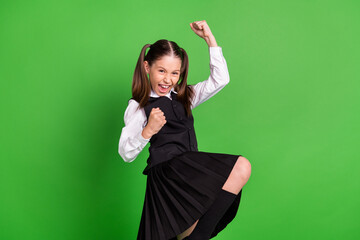 Photo of funny lucky school girl wear black white uniform rising fists smiling isolated green color background