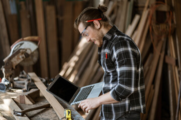 young hipster carpenter man holding computer laptop looking and checking wood in workshop ....