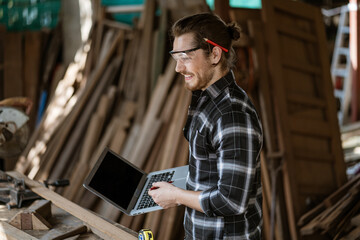 young hipster carpenter man holding computer laptop looking and checking wood in workshop ....