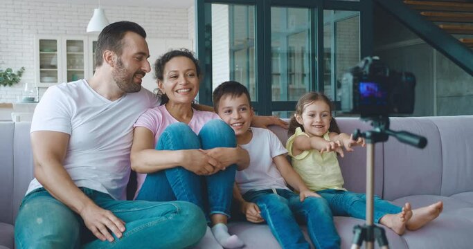 Woman sits next to her family on the couch, making video blog on professional video camera, actively gesticulates, referring to her husband and children, describes the new room by video recording