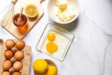 Baking ingredients on white table with copy space; top view. Eggs, flour, honey, lemon, cream, cream cheese and kitchen utensils on table.