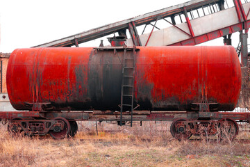 Fototapeta na wymiar a red shabby and rusty wagon from a freight train in cloudy weather in a field against the background of an abandoned factory