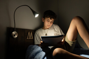 Boy in his room lying on his bed reading some notes at night with the illumination of his bedside lamp