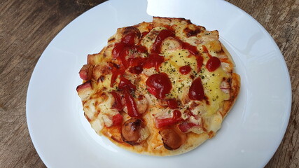 Homemade mini pizza with ham, mozzarella cheese, cheese and tomato sauce Sprinkle on thick pizza dough. Served in a round white plate on a wooden table. It is a favorite dish that children can make
