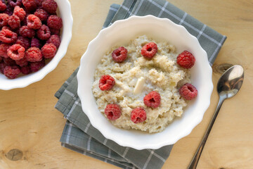 Milk barley porridge with butter and raspberries in white bowl on wooden background, top view