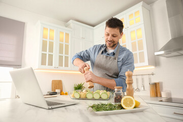 Man adding spices to dish while watching online cooking course via laptop in kitchen