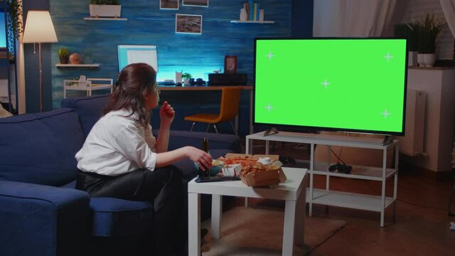 Woman looking at horizontal green screen on television while sitting on sofa eating chips. Young adult watching isolated background with chroma key and mockup template in living room after work.