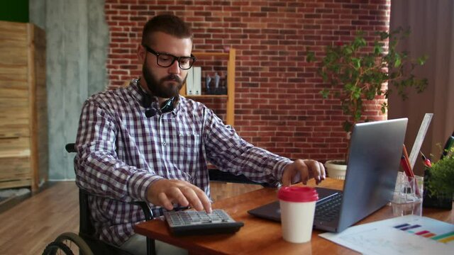 Businessman with glasses in wheelchair conducts budget calculations. Disabled hipster works on laptop using calculator, side view. Online business concept