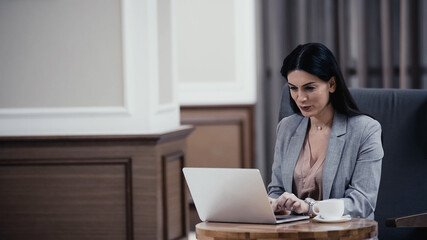 Obraz na płótnie Canvas brunette businesswoman using laptop near cup of coffee in lobby of restaurant