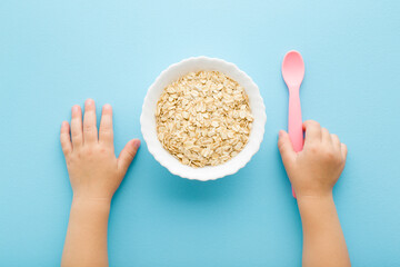 Baby hand holding pink plastic spoon. White bowl with dry rolled oat on light blue table...