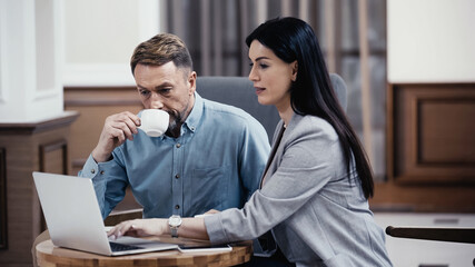 Businessman drinking coffee and looking at laptop near woman in lobby of restaurant