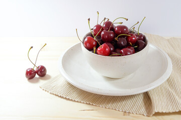 Organic sweet cherries in an off-white ceramic bowl and plate with a napkin on a light wooden table, fresh fruits as summer snack, copy space