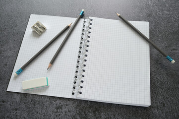 Working desktop with a blank open spiral notebook, pencils, sharpener and eraser on a dark gray table, mock up with copy space