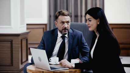 man in suit looking at cheerful businesswoman near laptop and cups on table