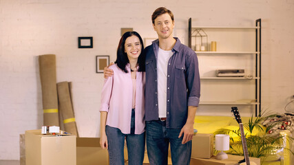 Fototapeta na wymiar cheerful man and woman hugging while standing near boxes in new home