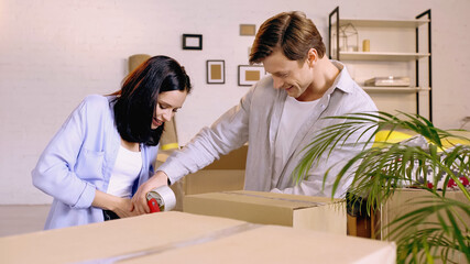 Obraz na płótnie Canvas joyful couple using scotch tape while packing box