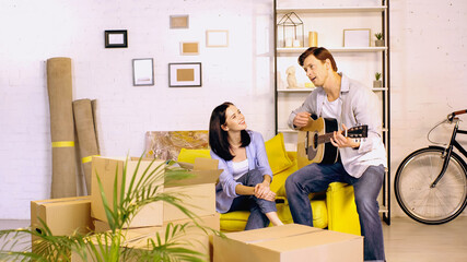 man playing acoustic guitar near happy woman sitting on couch in new home