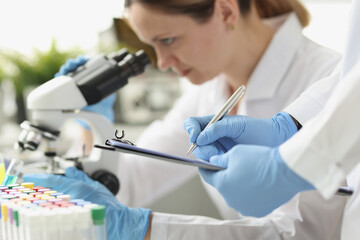 Woman chemist looking through microscope in laboratory. Assistant writing data to paper