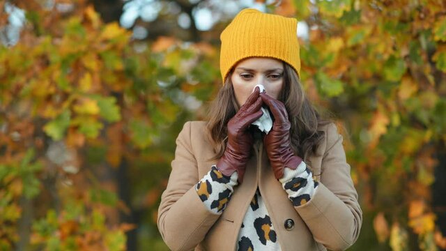 Hello october. young woman in beige coat and orange hat with napkin blowing nose outside in the city park in autumn. This video was made with PRORes 422 codec.
