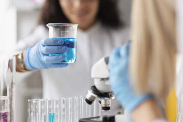 Fototapeta premium Woman chemist holding glass beaker with blue liquid in front of microscope in laboratory