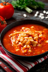 White bean soup with vegetables in a black bowl on a dark wooden background. Greek bean soup with tomatoes, carrots, celery, and onions. Fasolada