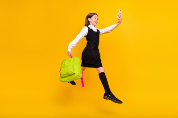 Full length body size photo schoolgirl jumping with schoolbag taking selfie isolated bright yellow color background