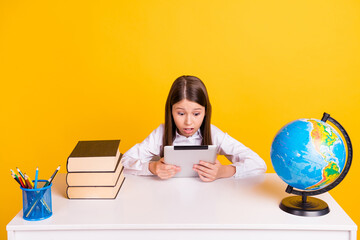 Photo portrait schoolgirl sitting at desk reading e-book on lesson shocked amazed isolated vivid yellow color background