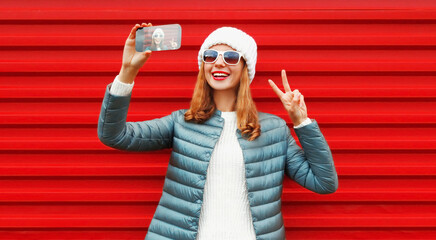 Portrait of cheerful happy laughing young woman taking selfie by smartphone wearing a white hat, jacket on red background