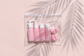 Feminine flatlay with travel size pink cosmetic bottles in bag on neutral pastel background. Palm leaves shadows. Minimalist skin or body care beauty products for vacation or journey. Top view.