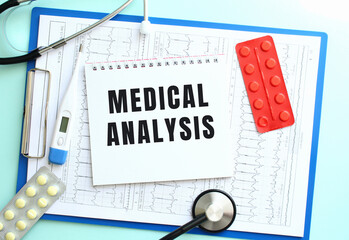 A notepad with the text MEDICAL ANALYSIS lies on a medical clipboard with a stethoscope and pills...