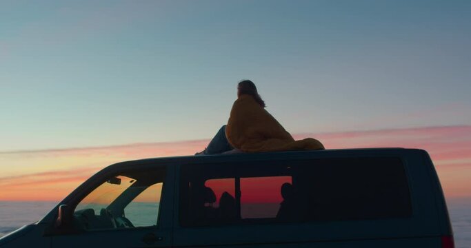 Cinematic shot of young woman cosy cuddled in yellow sleeping bag or blanket, sit on top of camper van. Young female traveller watch sunset from roof or camping van. Wanderlust van life concept